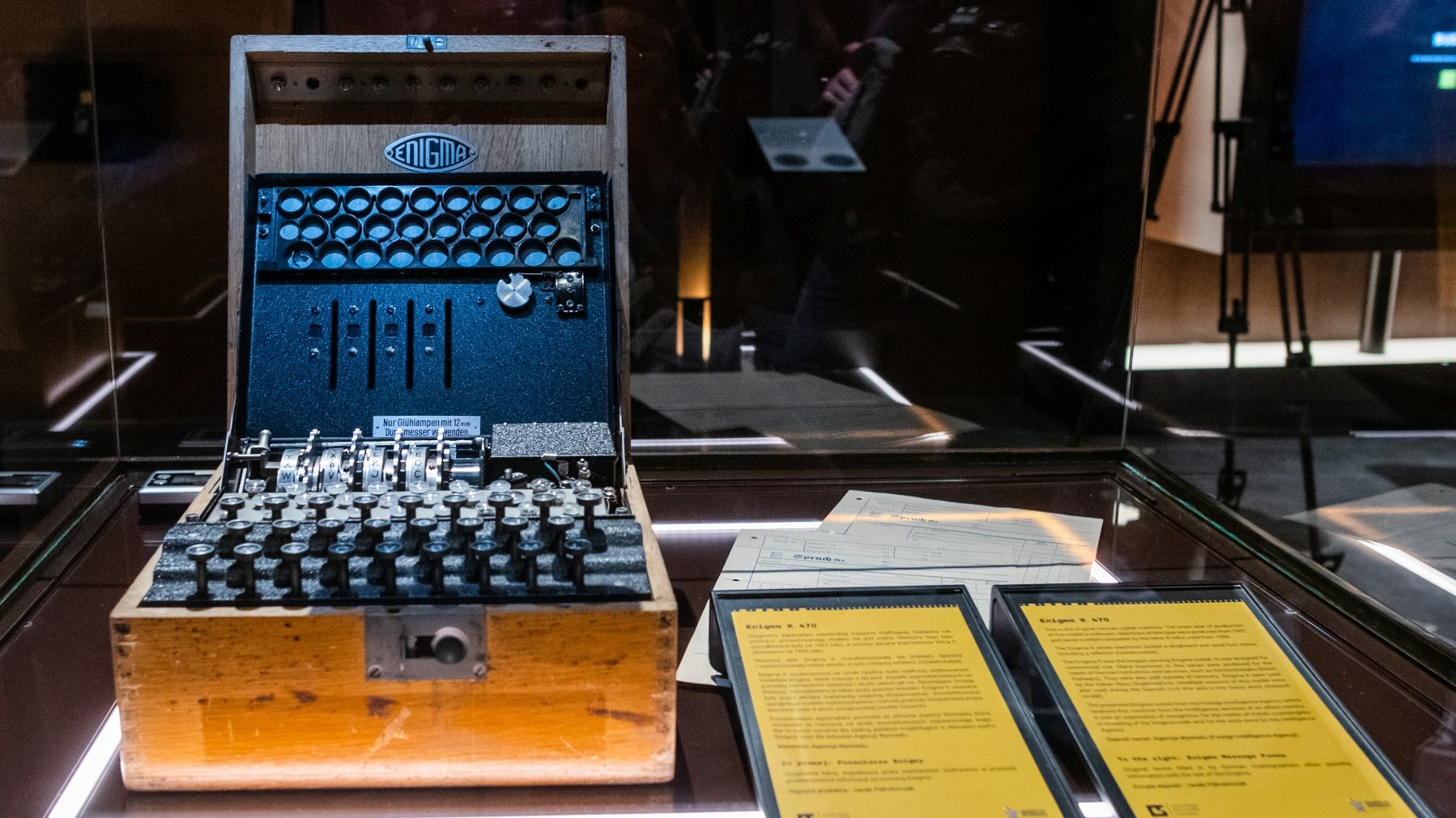 Zdjęcie przedstawia niemiecką maszynę szyfrującą Enigma umieszczoną w gablocie. Obok urządzenia widać dwie tabliczki informacyjne z opisem.