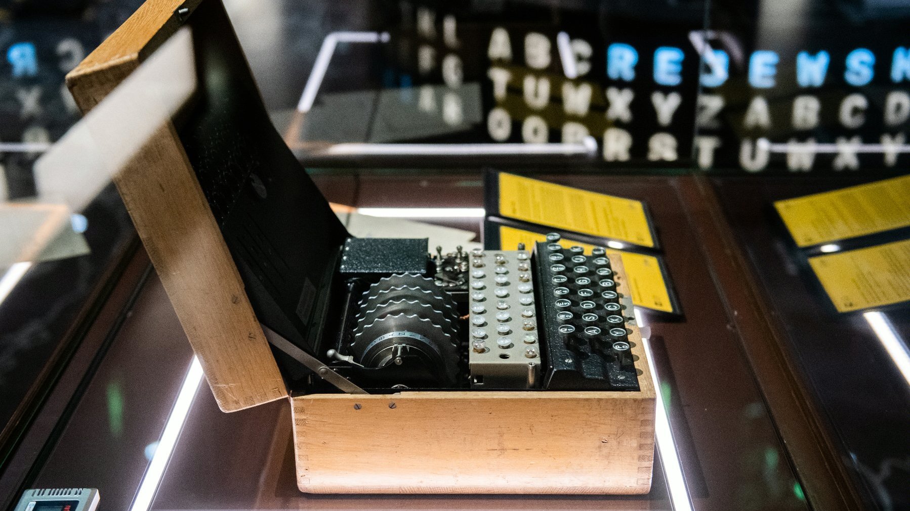 Zdjęcie przedstawia niemiecką maszynę szyfrującą Enigma umieszczoną w gablocie. Obok urządzenia widać dwie tabliczki informacyjne z opisem.