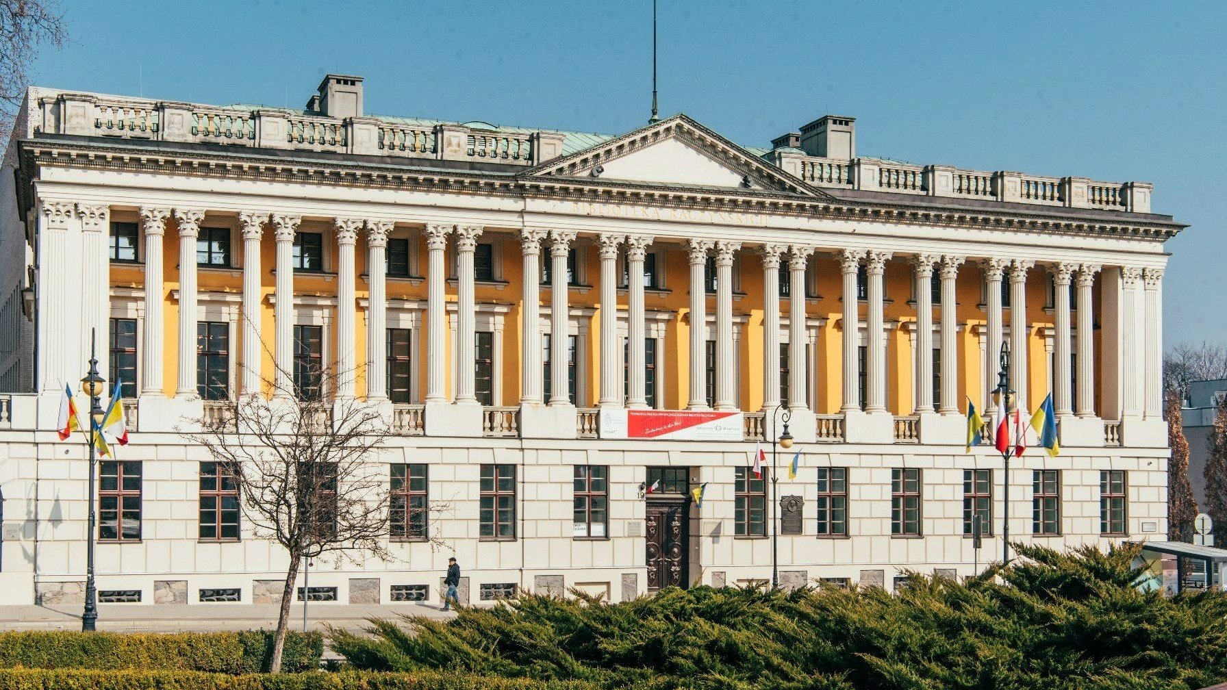 Na zdjęciu żółto-biały gmach biblioteki Raczyńskich, przed nim trawa i krzaki - grafika artykułu