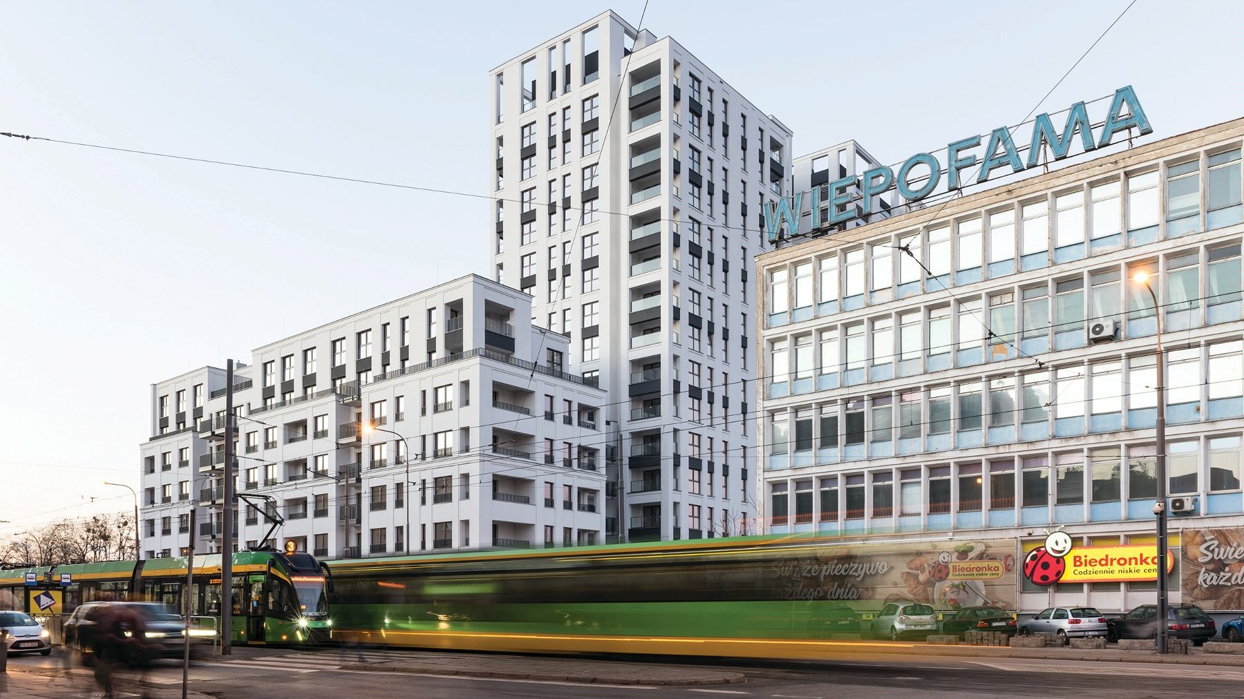 Zdjęcie przedstawia budynek "Famma". Na fotografii widaać wyraźny podział bryły na dwie części - niską, uzupełniającą pierzeję, oraz kilkunastopiętrową, wysoką. Na pierwszym planie widać przejeżdzające tramwaje.