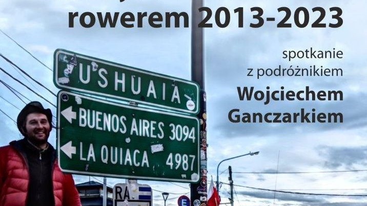 Zdjęcie na spotkanie z podróżnikiem Wojciechem Ganczarkiem. Uśmiechnięty mężczyzna w czerwonej kurtce stoi przy rowerze obwieszonym podróżniczym ekwipunkiem. W tle szare niebo, linie napięcia, auta, ulica w Ameryce Południowej.