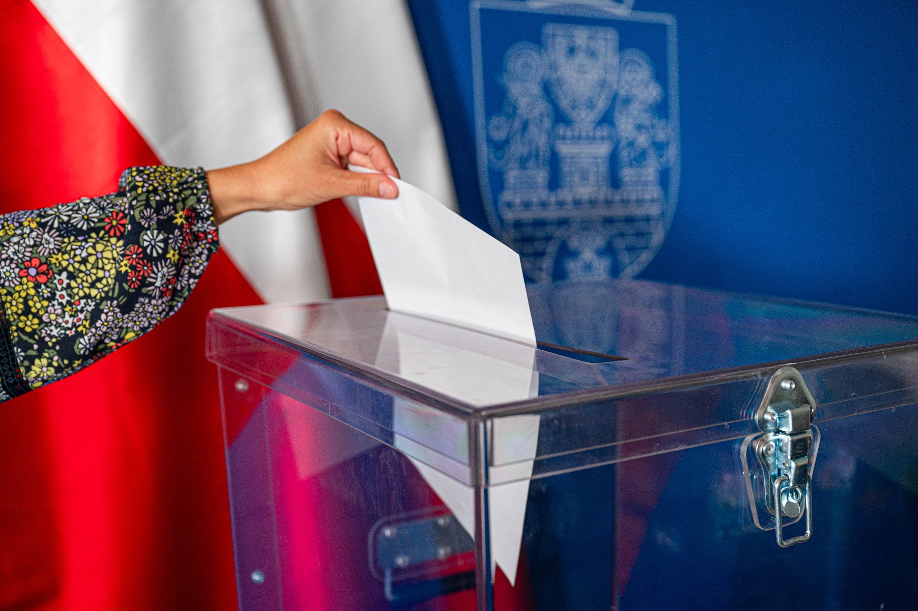 Na zdjęciu widać urnę wyborczą oraz rękę, która wrzuca do urny kartę do głosowania. W tle herb Poznania oraz flagi Polski. - grafika artykułu