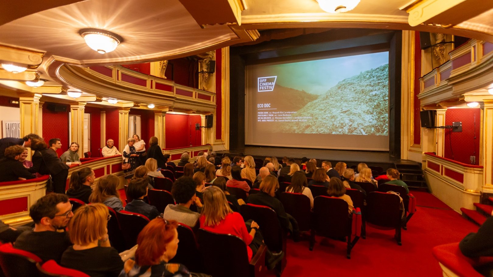 Na zdjęciu sala teatralna wypełniona ludźmi, na scenie ekran