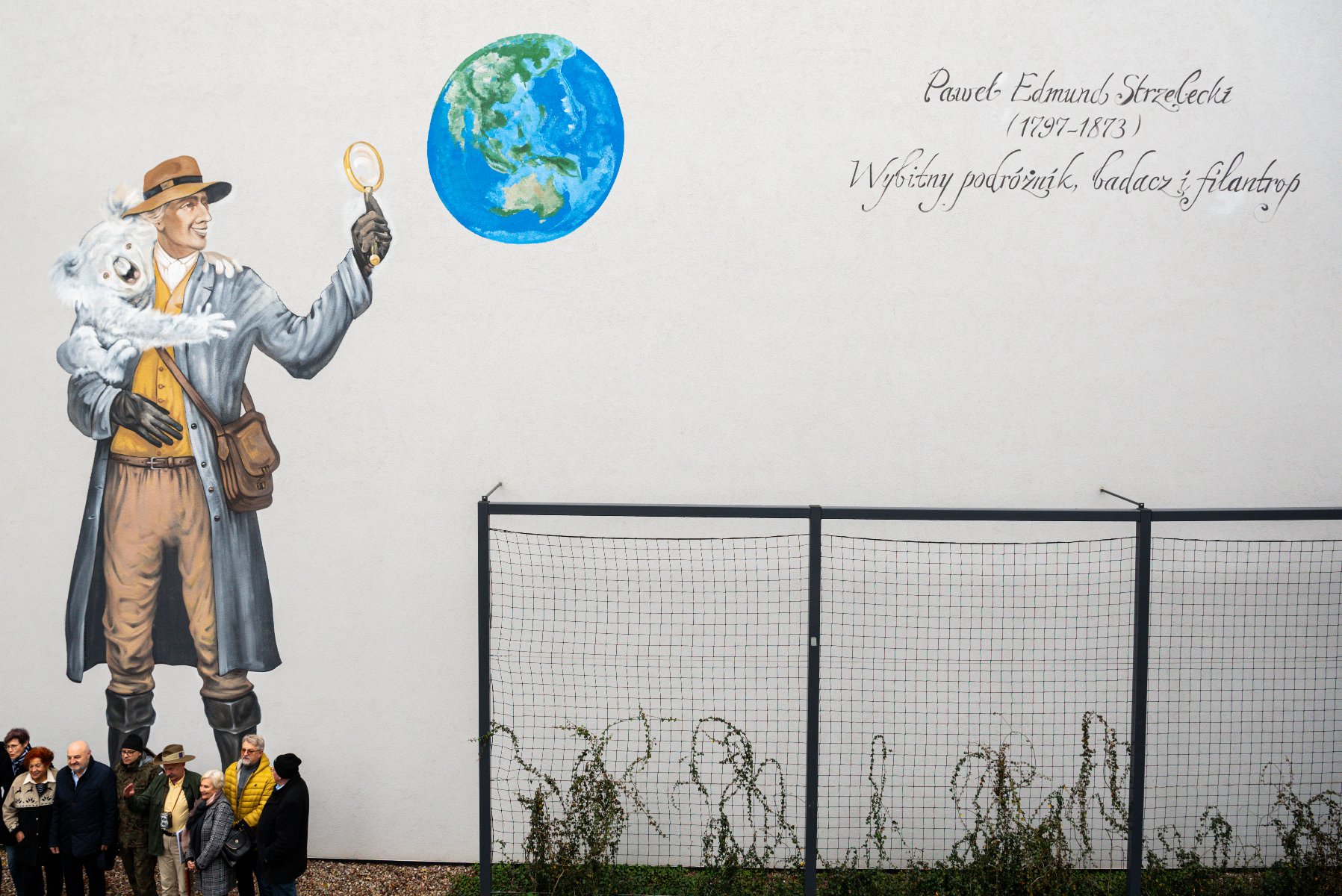 Zdjęcia z odsłonięcia muralu z Pawłem Edmundem Strzeleckim - na muralu mężczyzna z koalą na ramieniu, mężczyzna trzyma w ręku lupę, przez którą patrzy na kulę ziemską. Dookoła zgromadzeni ludzie - grafika artykułu