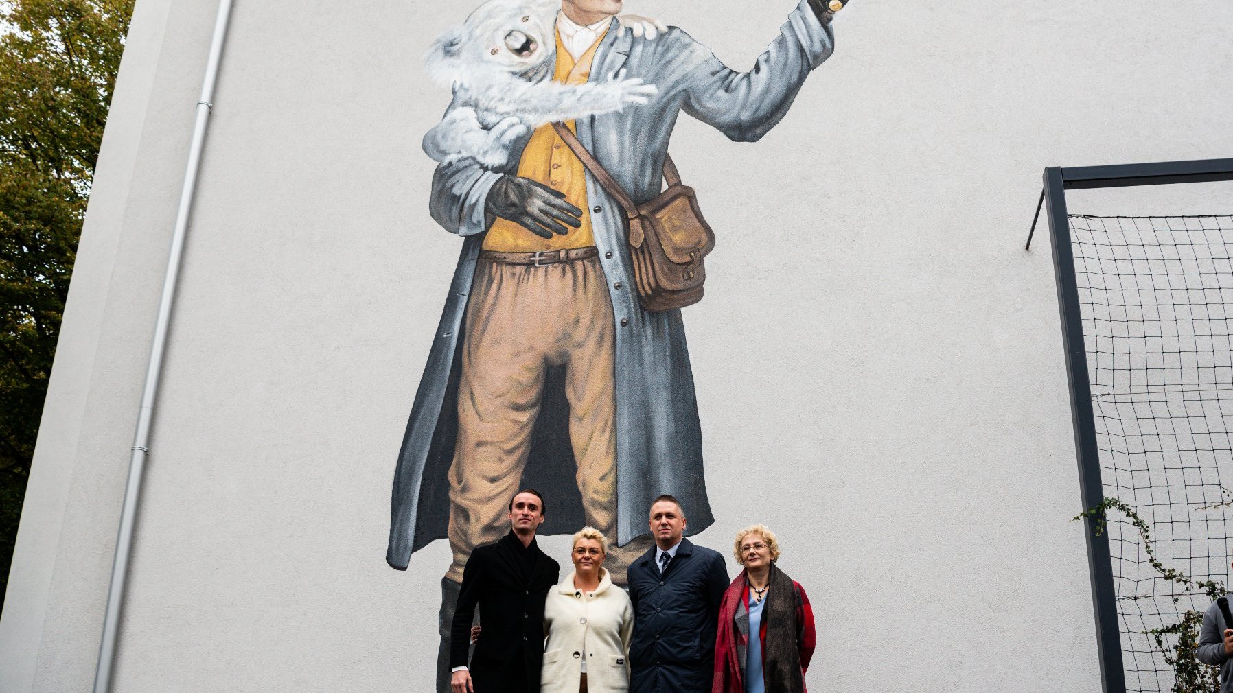 Zdjęcia z odsłonięcia muralu z Pawłem Edmundem Strzeleckim - na muralu mężczyzna z koalą na ramieniu, mężczyzna trzyma w ręku lupę, przez którą patrzy na kulę ziemską. Dookoła zgromadzeni ludzie