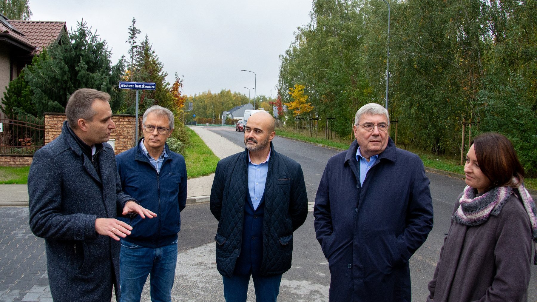 Grupa osób rozmawia ze sobą. W tle ul. Iwaszekiewicza - jezdnia, zabudowania, zieleń.