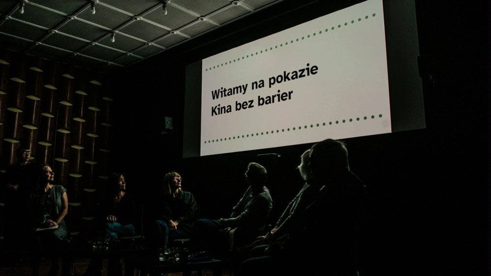Na zdjęciu widać grupę osób siedzącą na krzesłach w zaciemnionej sali, za plecami mają ekran, na którym wyświetlana jest grafika z napisem: Witamy na pokazie Kina bez barier.