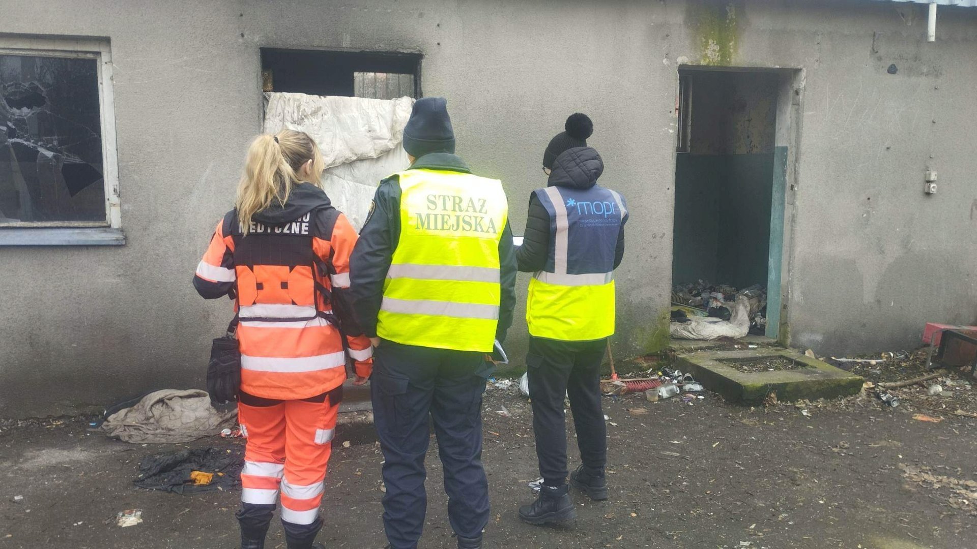 Na zdjęciu trzy osoby: pracownik MOPR, strażnik miejski i ratowniczka medyczna stoją przed opuszczonym budynkiem