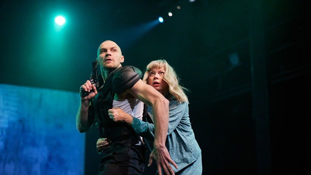 Na zdjęciu dwójka ludzi na scenie, kobieta przestraszona chowa się za mężczyzną, który trzyma pistolet