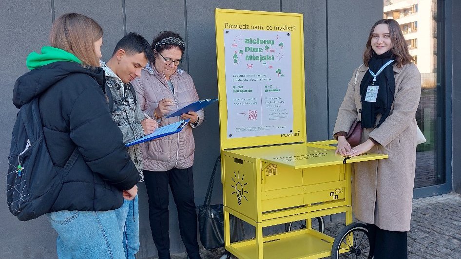 Na zdjęciu czwórka ludzi przez żółtym wózkiem a plakatami kampanii