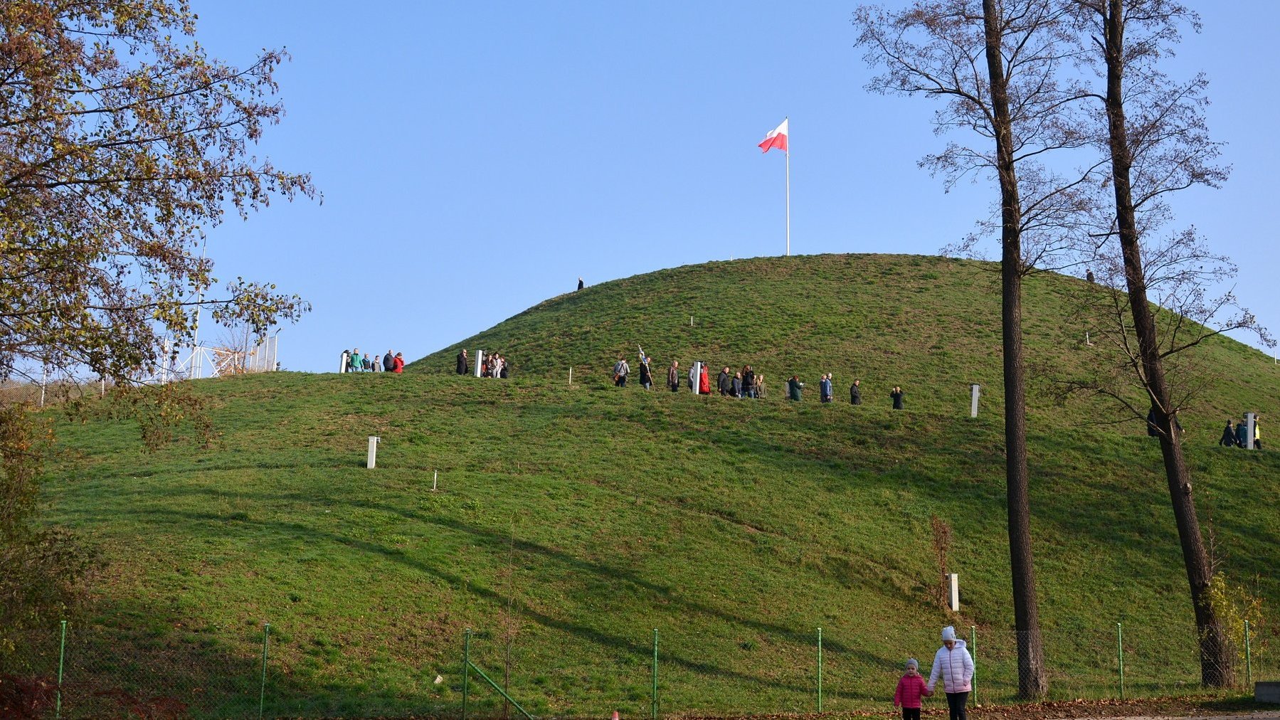 Galeria zdjęć przedstawia kopiec ziemny porośnięty trawą, na jego szczycie powiewa flaga Polski.