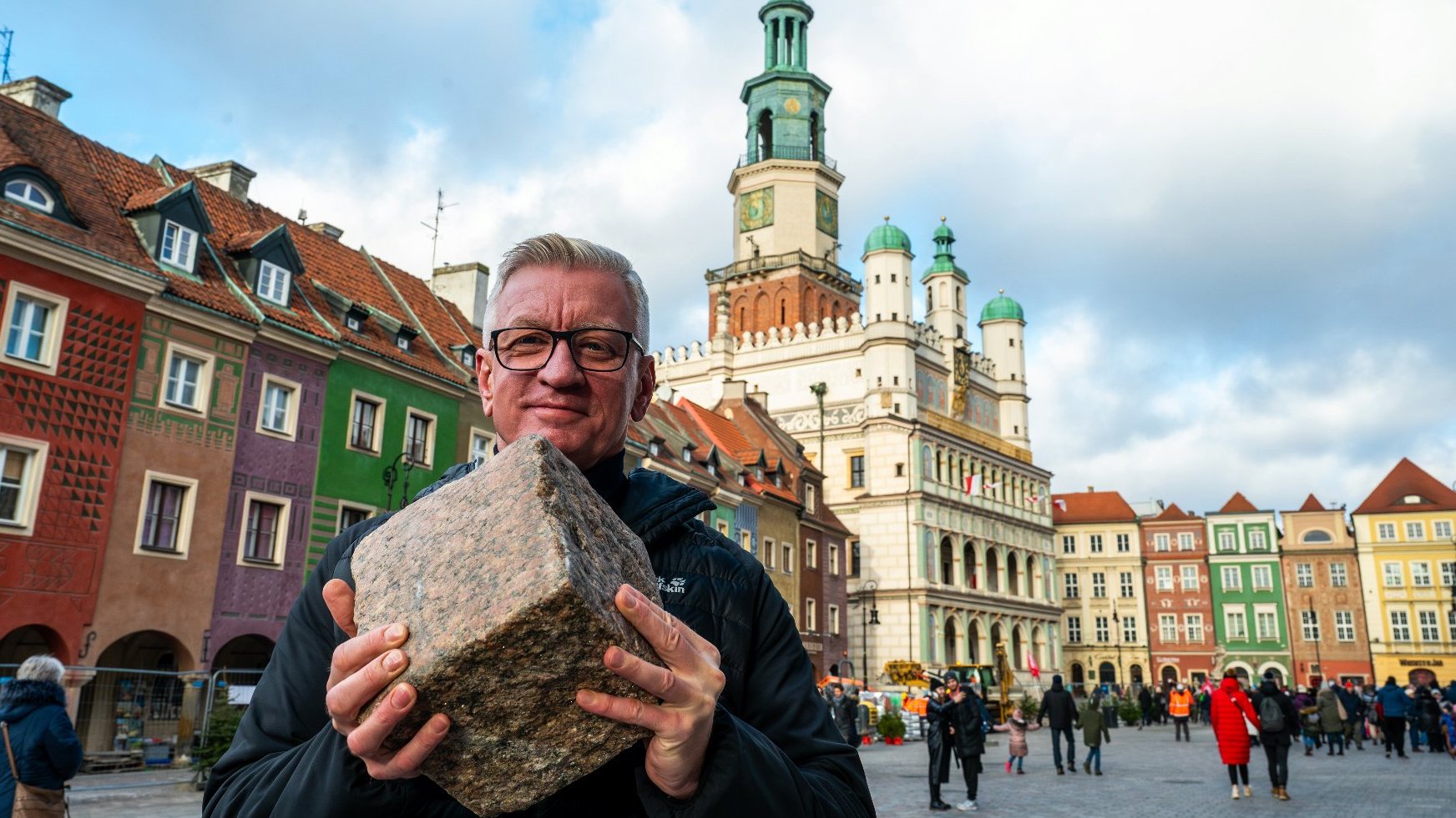 Na pierwszym planie Jacek Jaśkowiak trzymający w dłoniach kamienną kostkę. W tle ratusz i domki budnicze.