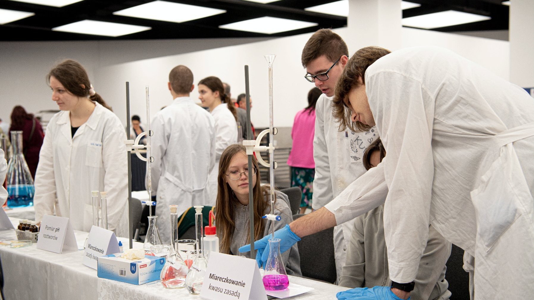 Zdjęcie przedstawia młodzież w białych fartuchach za stołem laboratoryjnym.