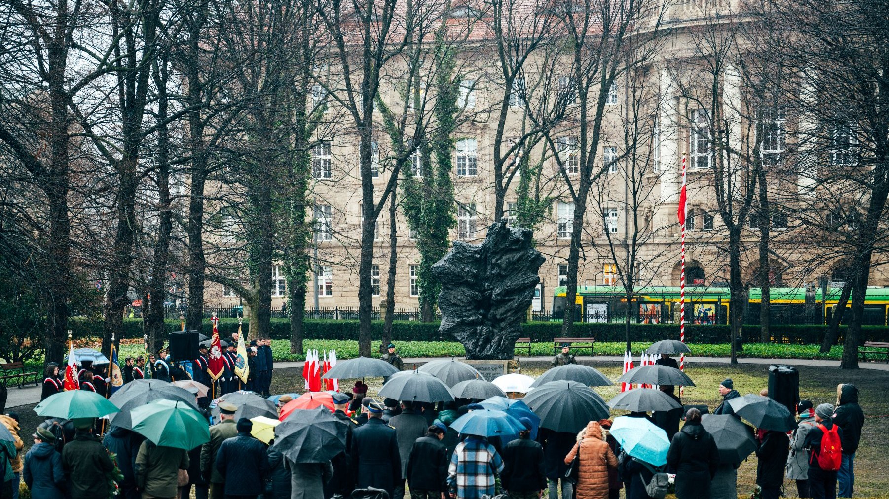 Na zdjęciu pomnik, pod nim zgromadzeni ludzie z otwartymi parasolami