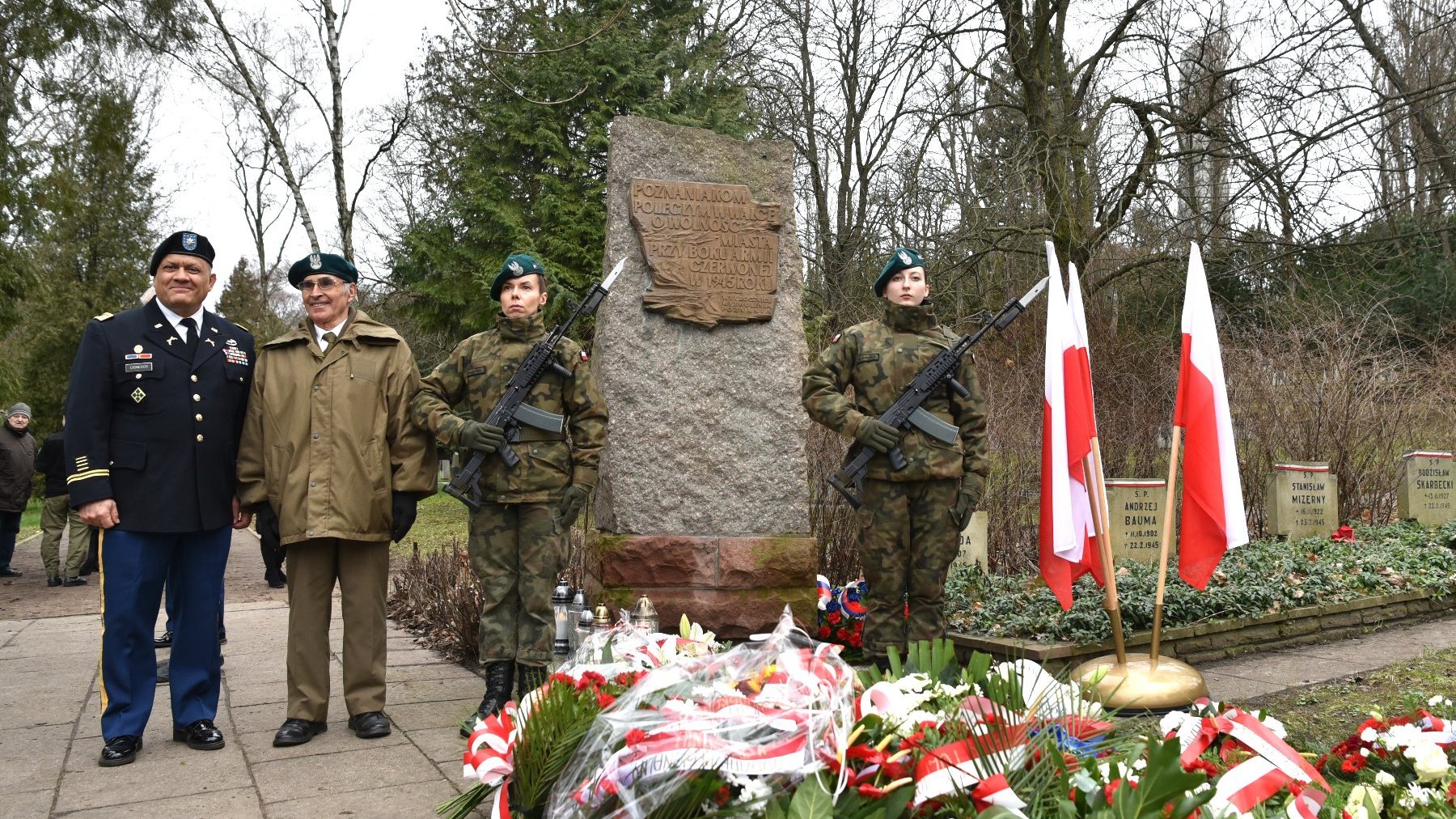 Zdjęcie przedstawia pomnik pod kórym leżą kwiaty. Przed pomnikiem stoją żołnierze.