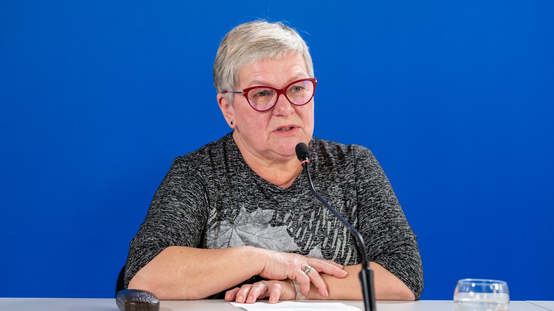 Na zdjeciu kobieta w okularach siedząca za stołem konferencyjnym, mówiąca coś do mikrofonu