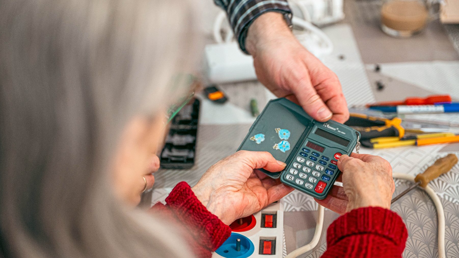 Na zdjęciu dłonie starszej kobiety i mężczyzny trzymających mały kalkulator