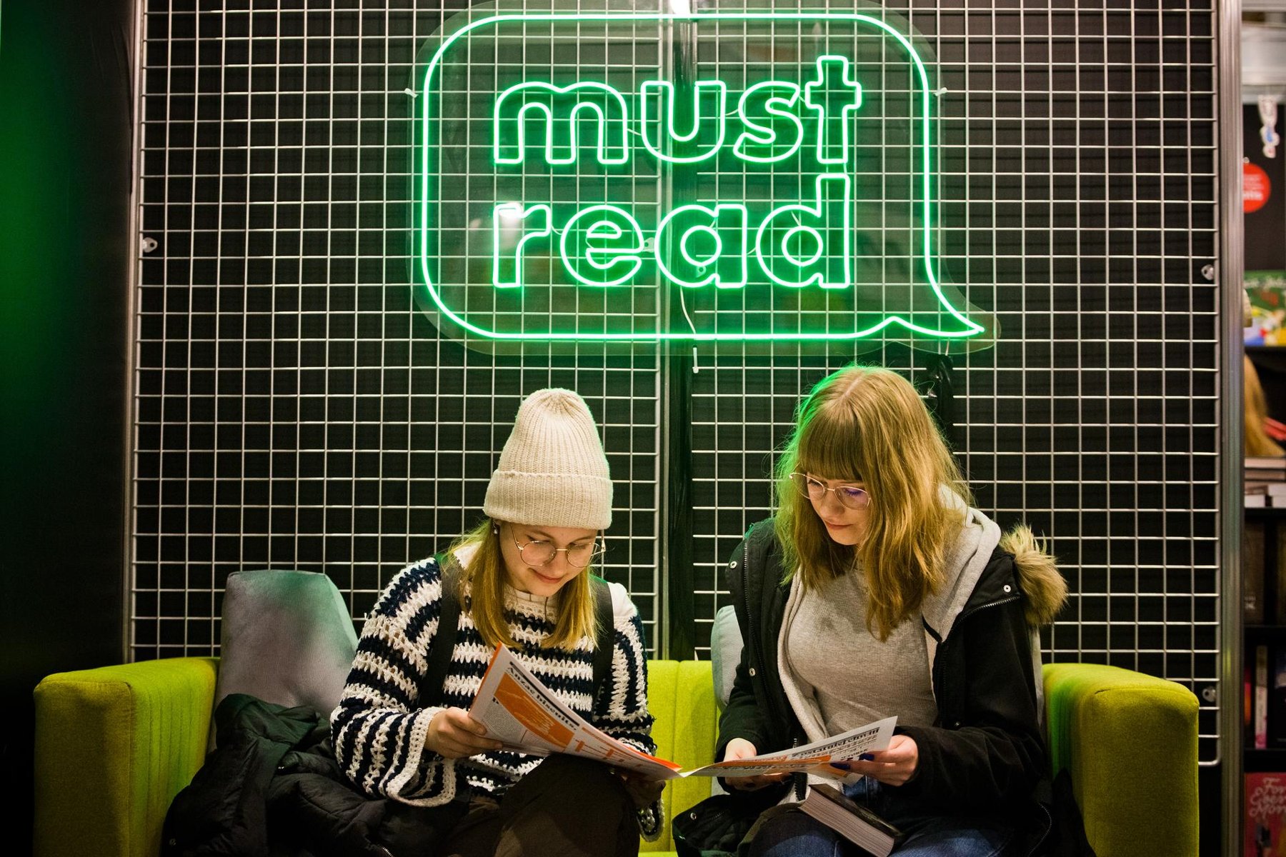 Na zdjęciu dwie kobiety czytające gazetę pod neonem z napisem: must read - grafika artykułu