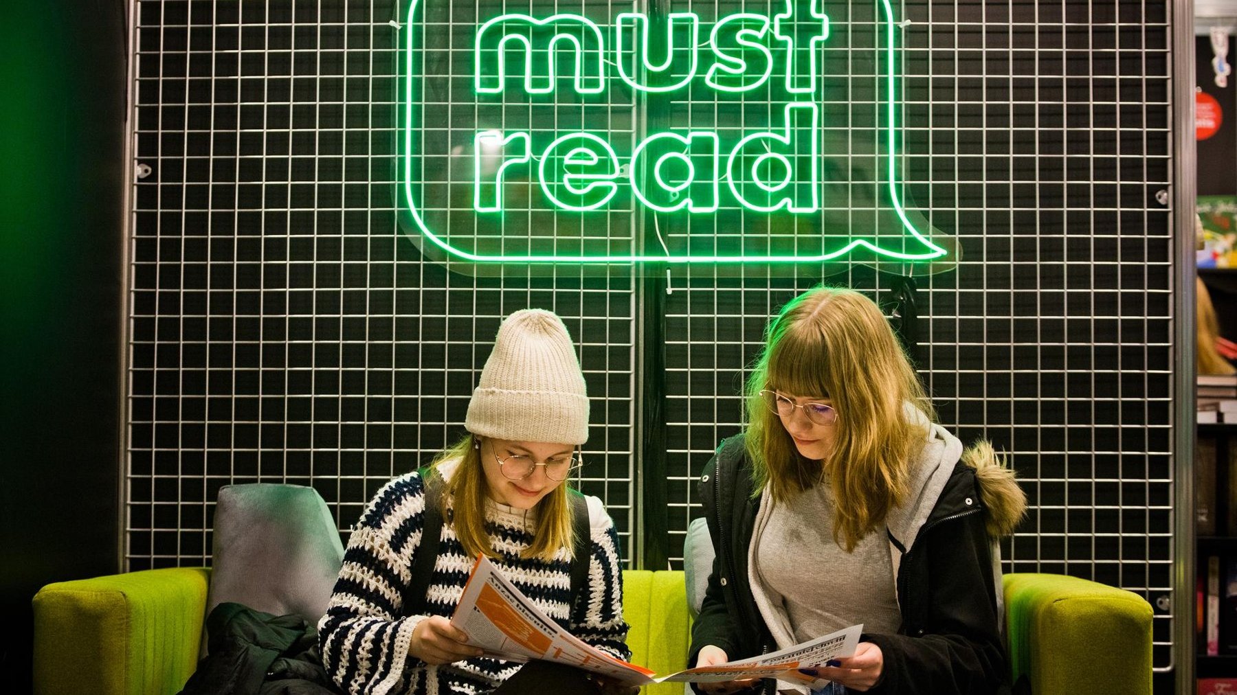 Na zdjęciu dwie kobiety czytające gazetę pod neonem z napisem: must read