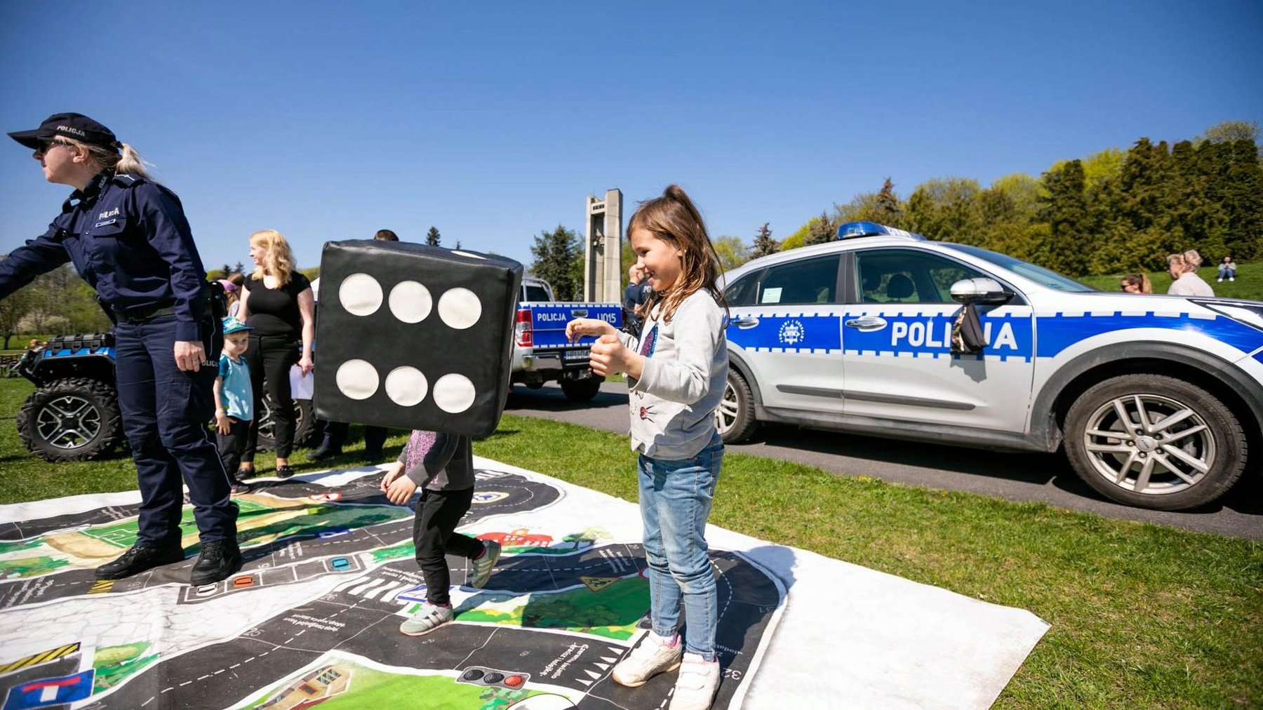 Na zdjęciu bawiące się dzieci, w tle radiowóz policji