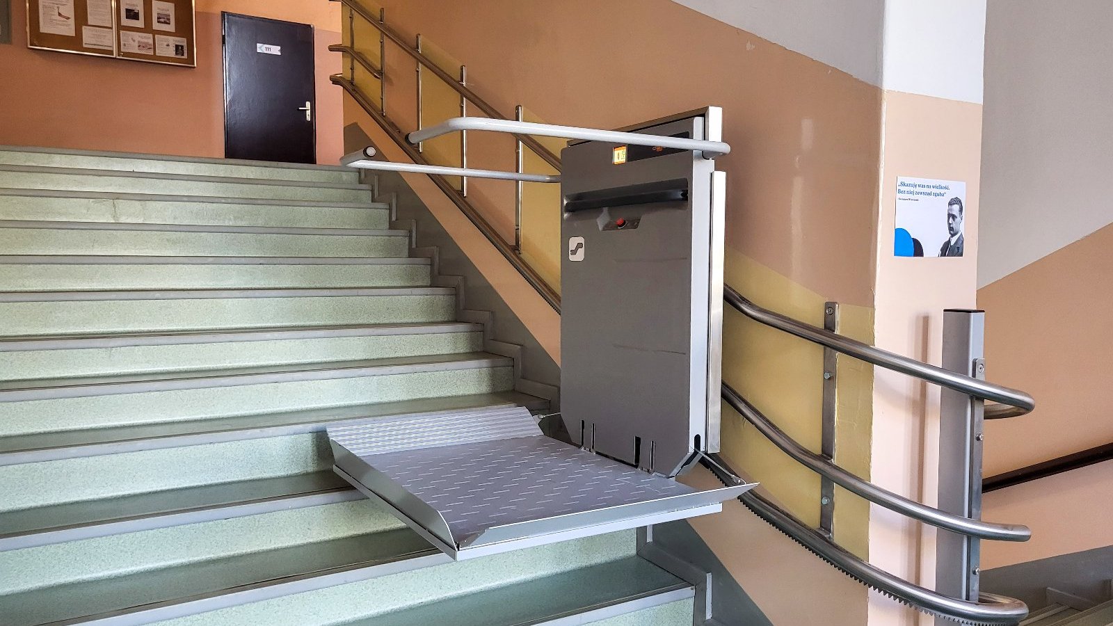Zdjęcie przedstawia windę dla wóźków zamontowaną nad schodami.