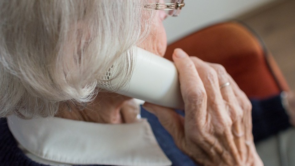 Na zdjęciu kobieta trzymająca słuchawkę telefonu przy uchu, siedząca tyłem do obiektywu