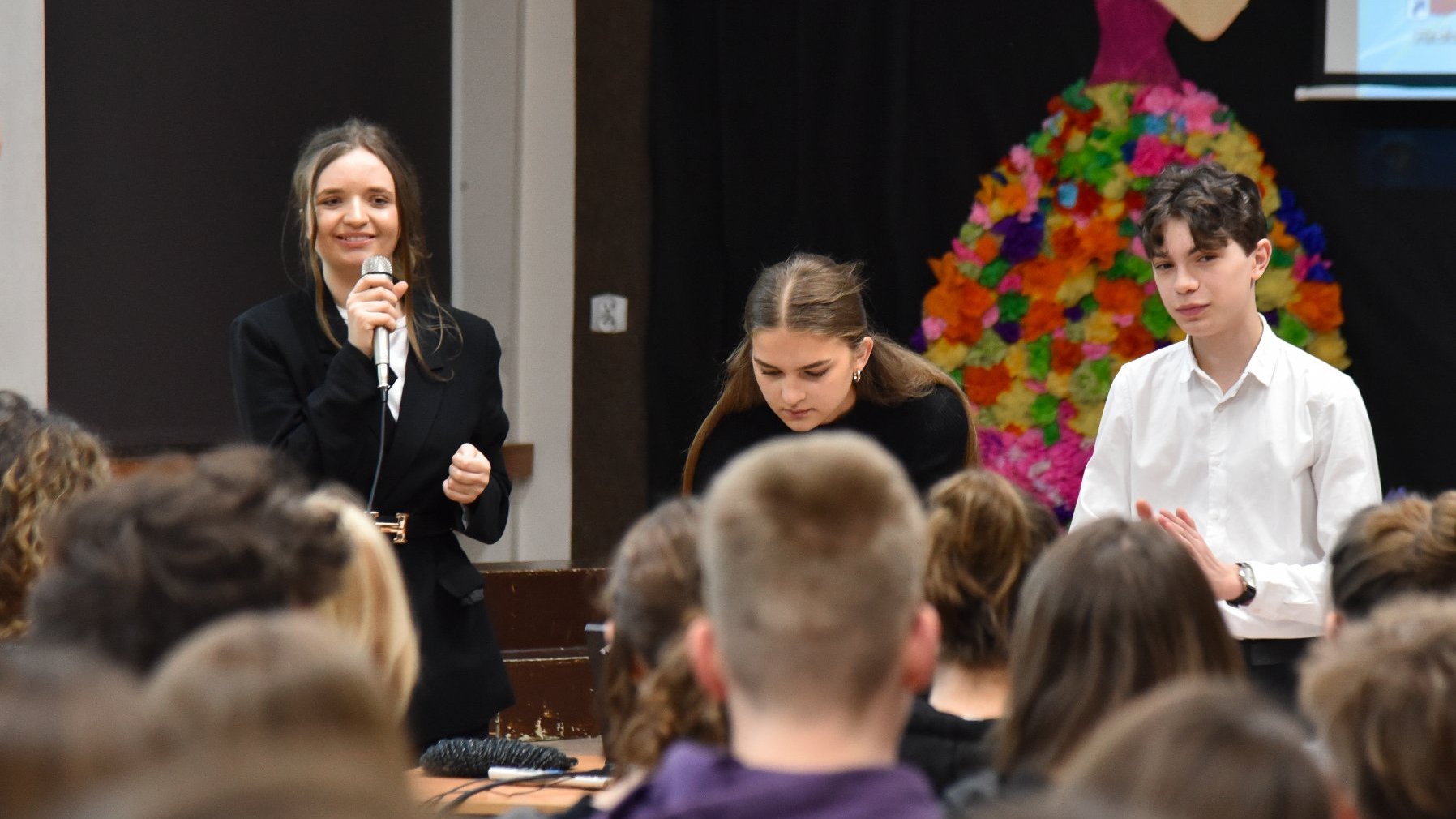 Na zdjęciu troje uczniów podczas prezentacji, jedna z dziewczyn trzyma w ręku mikrofon