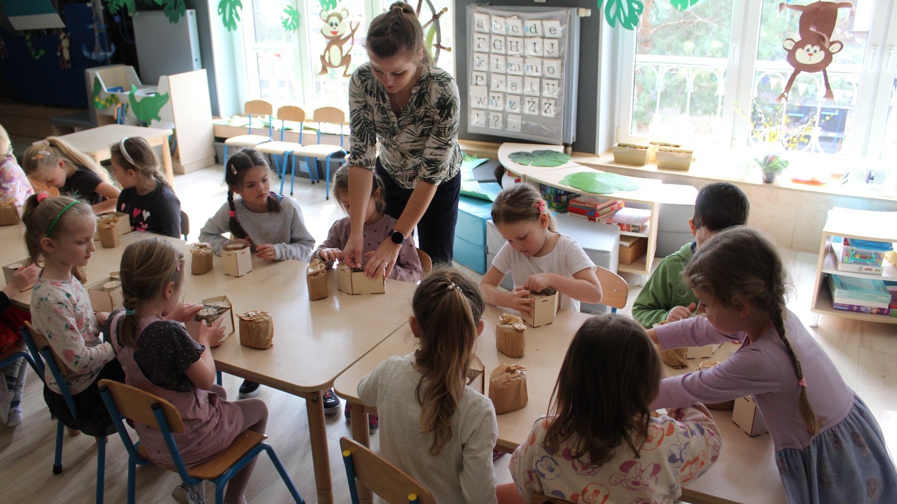 Zdjęcie przedstawia grupę dzieci przy stole podczas zajęć manualnych oraz kobietę prowadzącą lekcję.