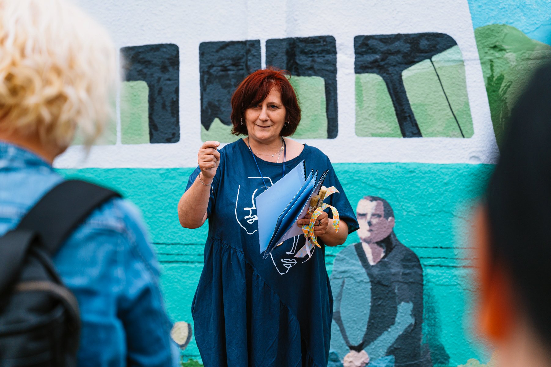 Na zdjęciu kobieta przed muralem, gestykuluje - grafika artykułu