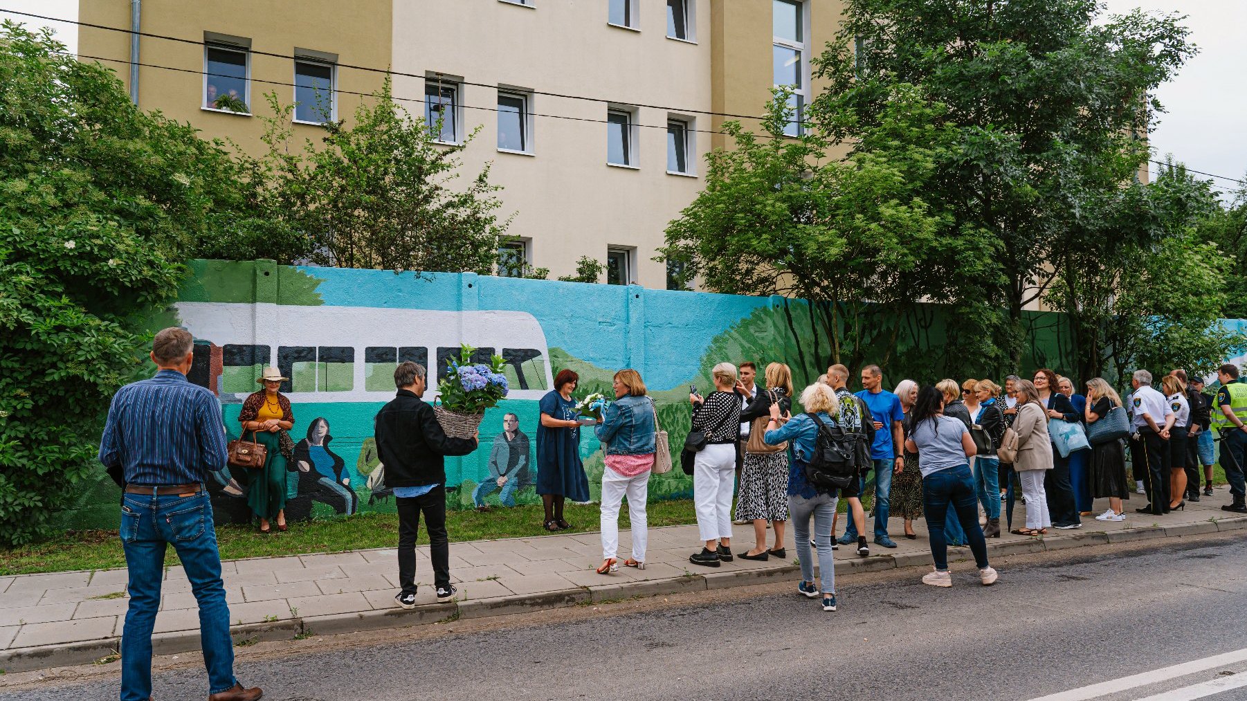 Na zdjęciu grupa ludzi zebrana przed murem, na murze mural