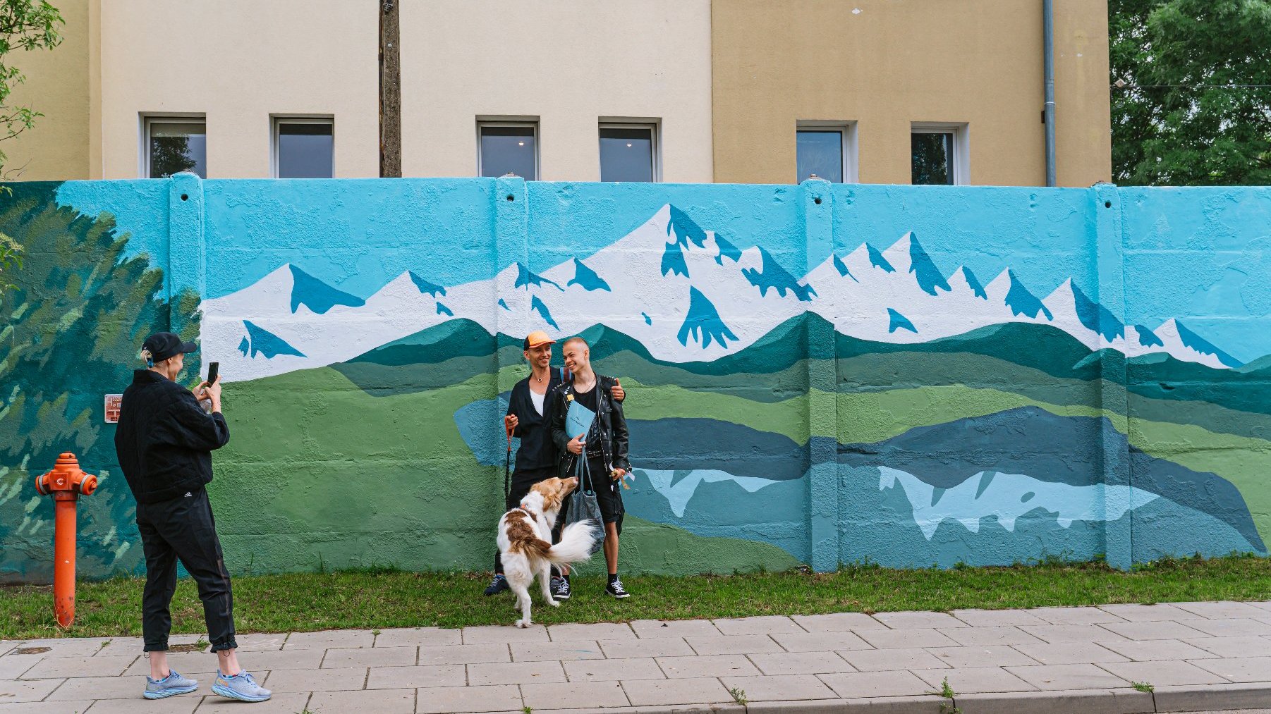 Na zdjęciu mural, przed nim ktoś robi zdjęcie dwóm osobom i psu