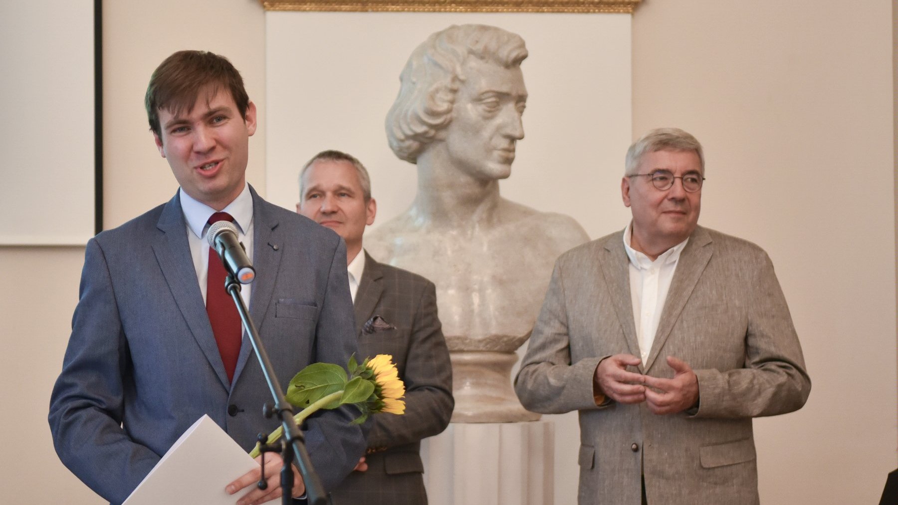 Na zdjęciu młody mężczyzna mówiący coś do mikrofonu, za ni9m dwóch innych mężczyzn