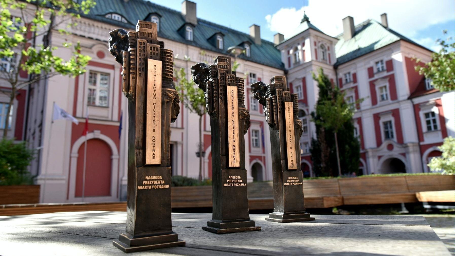 Zdjęcie przedstawia trzy statuetki Nagrody Prezydenta Miasta Poznania Architectus civitatis nostrae na tle budynku urzędu miasta przy pl. Kolegiackim.