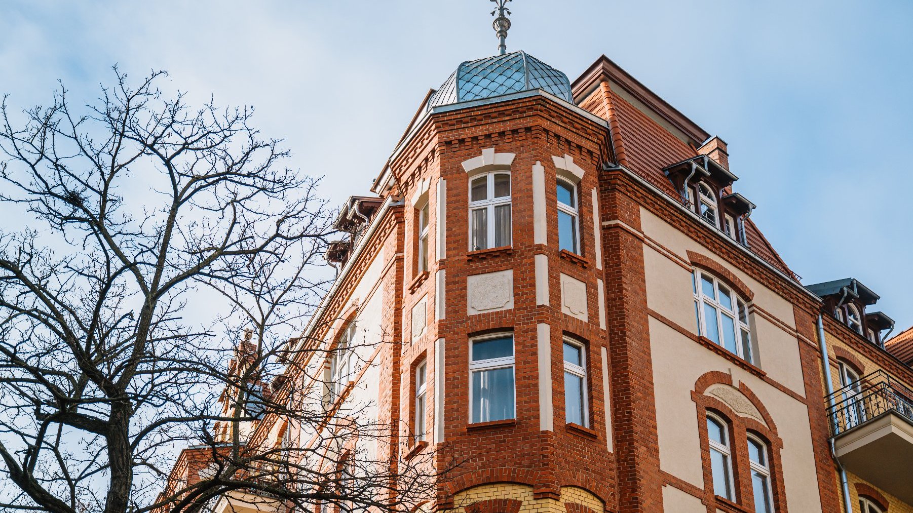 Budynkek mieszkalny o wielokondygnacyjnym stylu architektonicznym. Na zdjęciu widać połączenie czerwonej cegły z jasnym tynkiem oraz ozdobne detale wokół okien i linii dachowej. Dach ma stromy spadek z lukarnami i małą kopułą u góry. Widać kilka balkonów z kutymi żelaznymi balustradami. Na pierwszym planie znajduje się drzewo bez liści.