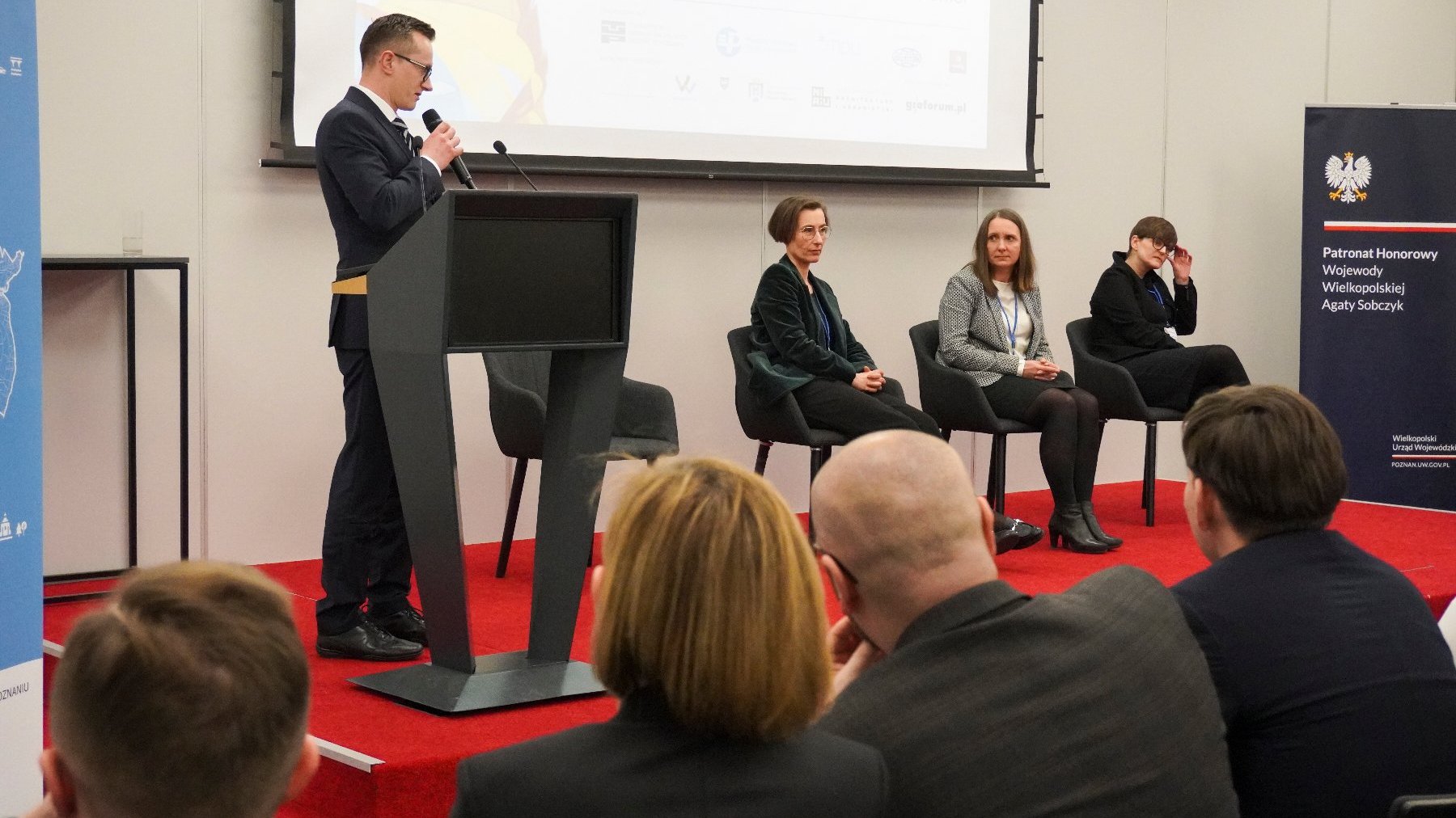 Wystąpienie na konferencji lub seminarium. Osoba przy mównicy mówi do mikrofonu, za nią siedzi trzech uczestników na scenie, słuchających uważnie. Za mównicą znajduje się duży ekran z tekstem i logami.