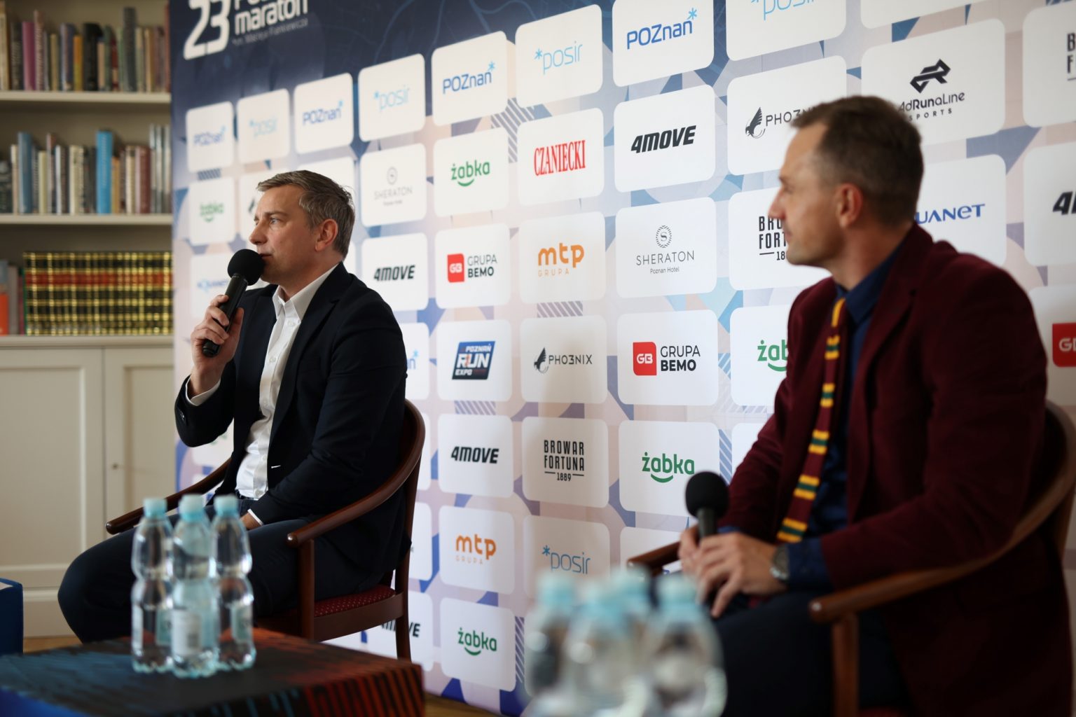 Dyrektor 23. Poznań Maratonu przemawia podczas konferencji prasowej poprzedzającej imprezę, fot. POSiR - 2 mężczyzn siedzi z mikrofonami na tle ścianki, jeden z nich przemawia - grafika artykułu