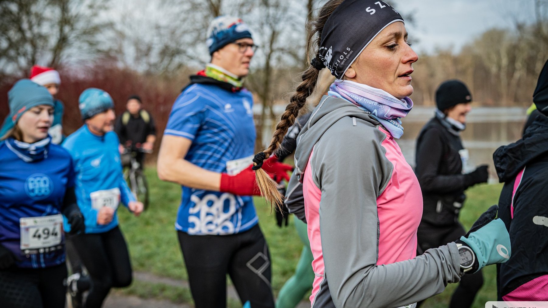 Bieg City Trail w Poznaniu, fot. Piotr Oleszak - na pierwszym planie biegaczka w szaro-różowym stroju, rękawiczkach i opasce. W tle inni biegacze w niebieskich i czarnych strojach w trakcie biegu, a także drzewa i krzewy oraz jezioro