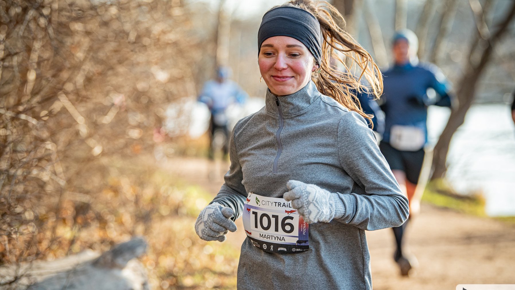 Bieg City Trail w Poznaniu, fot. Piotr Oleszak - na pierwszym planie uśmiechnięta biegaczka w szarym stroju, rękawiczkach i opasce. W tle kilku biegaczy pokonuje dystans w słonecznej pogodzie w lesie i ścieżce nad jeziorem