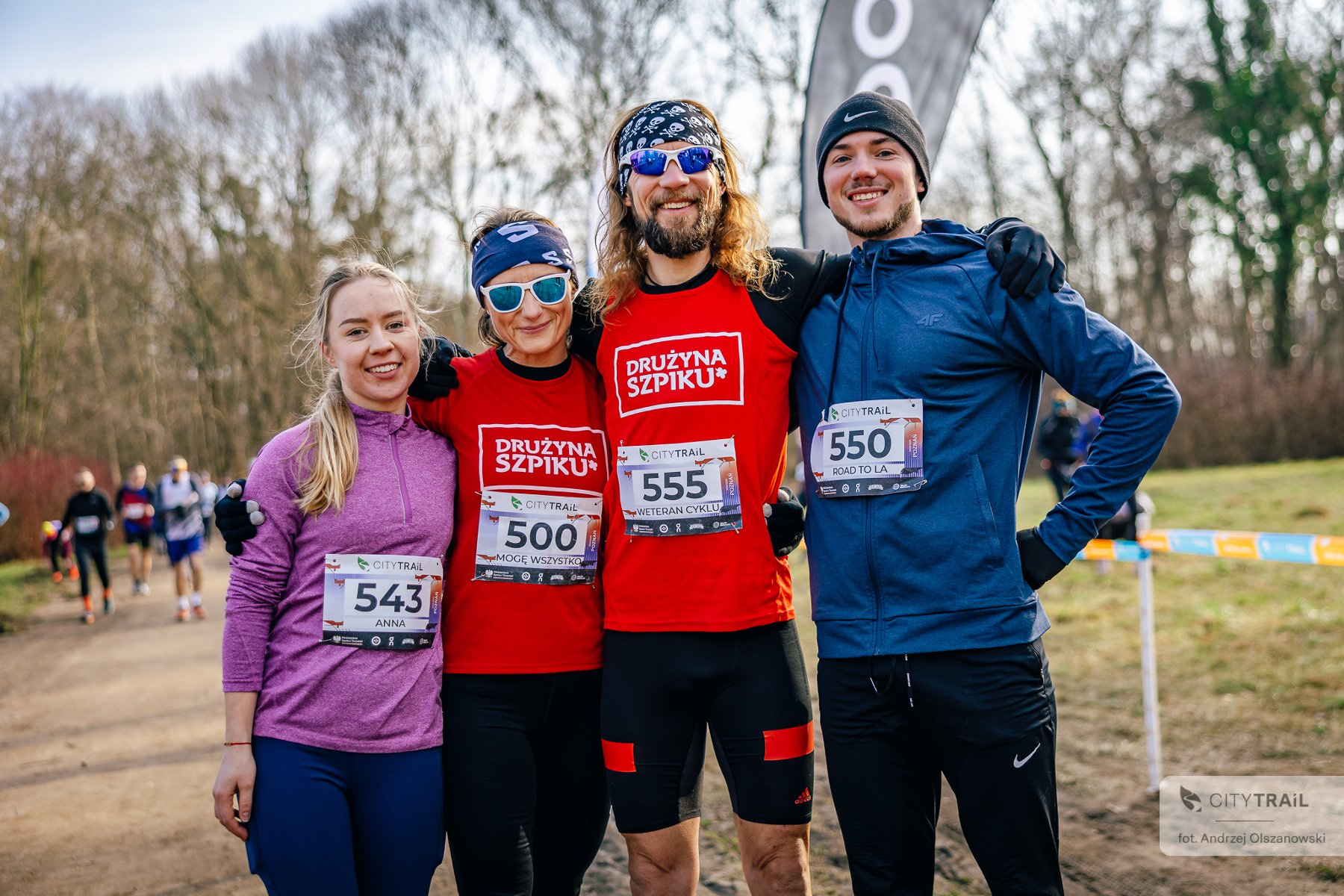 Bieg City Trail w Poznaniu, fot. Andrzej Olszanowski - 4 biegaczy stoi uśmiechniętych przed biegiem na trasie, 2 mężczyzn orz 2 kobiety. Kobieta po lewej w różowej bluzie, potem kobieta i mężczyzna w czerwonej koszulce "Drużyna Szpiku" i okularach przeciwsłonecznych, a także mężczyzna w czapce zimowej i niebieskiej bluzie. - grafika artykułu