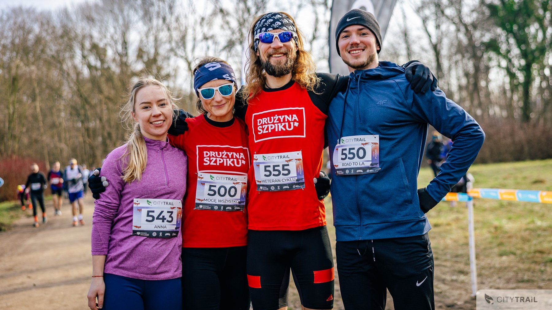 Bieg City Trail w Poznaniu, fot. Andrzej Olszanowski - 4 biegaczy stoi uśmiechniętych przed biegiem na trasie, 2 mężczyzn orz 2 kobiety. Kobieta po lewej w różowej bluzie, potem kobieta i mężczyzna w czerwonej koszulce "Drużyna Szpiku" i okularach przeciwsłonecznych, a także mężczyzna w czapce zimowej i niebieskiej bluzie.