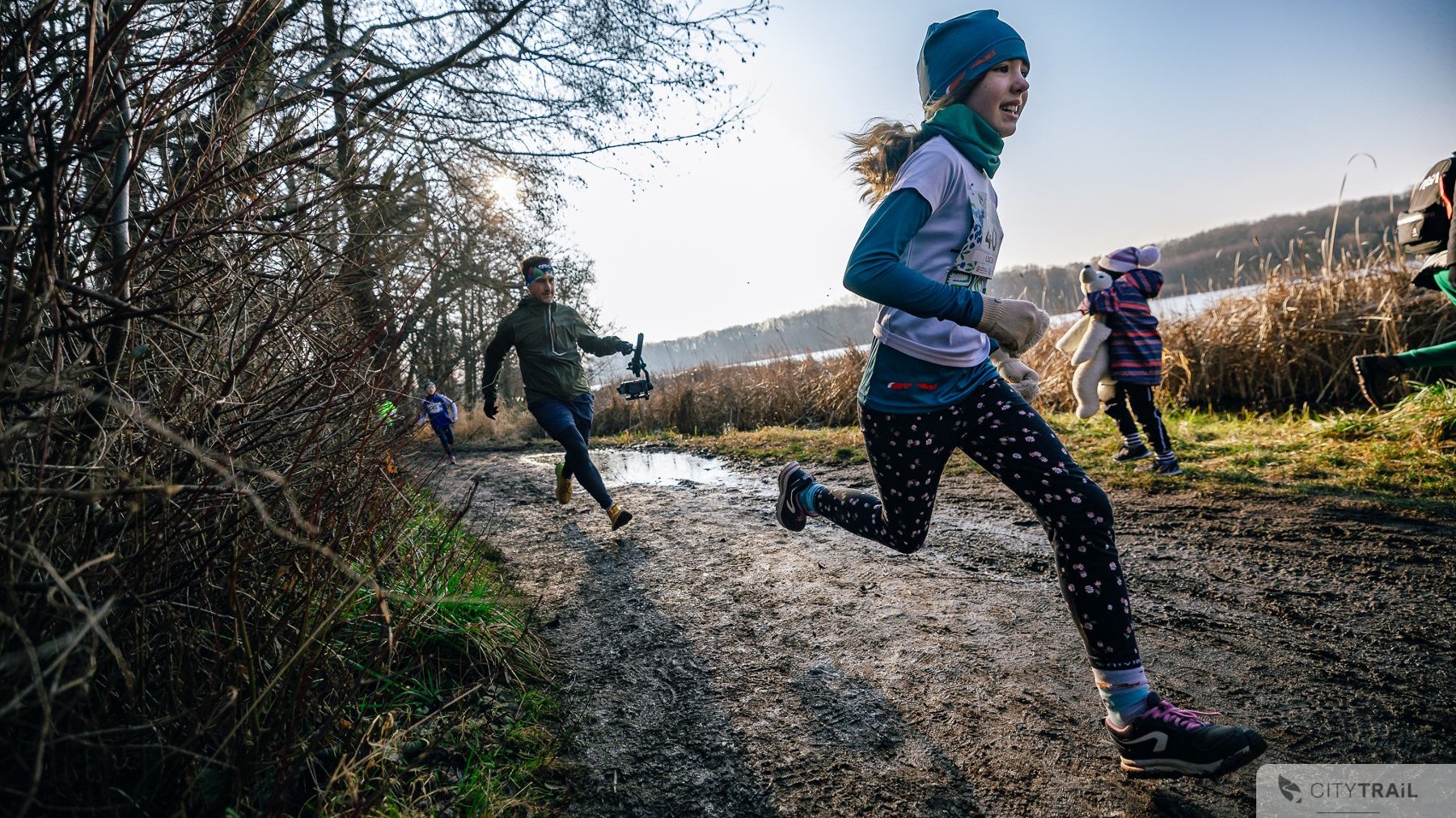 Bieg City Trail w Poznaniu, fot. Andrzej Olszanowski - na pierwszym planie biegnąca dziewczynka w błocie. W tle widać kolejne osoby biegnące, w tym mężczyzna w zielonej bluzie.