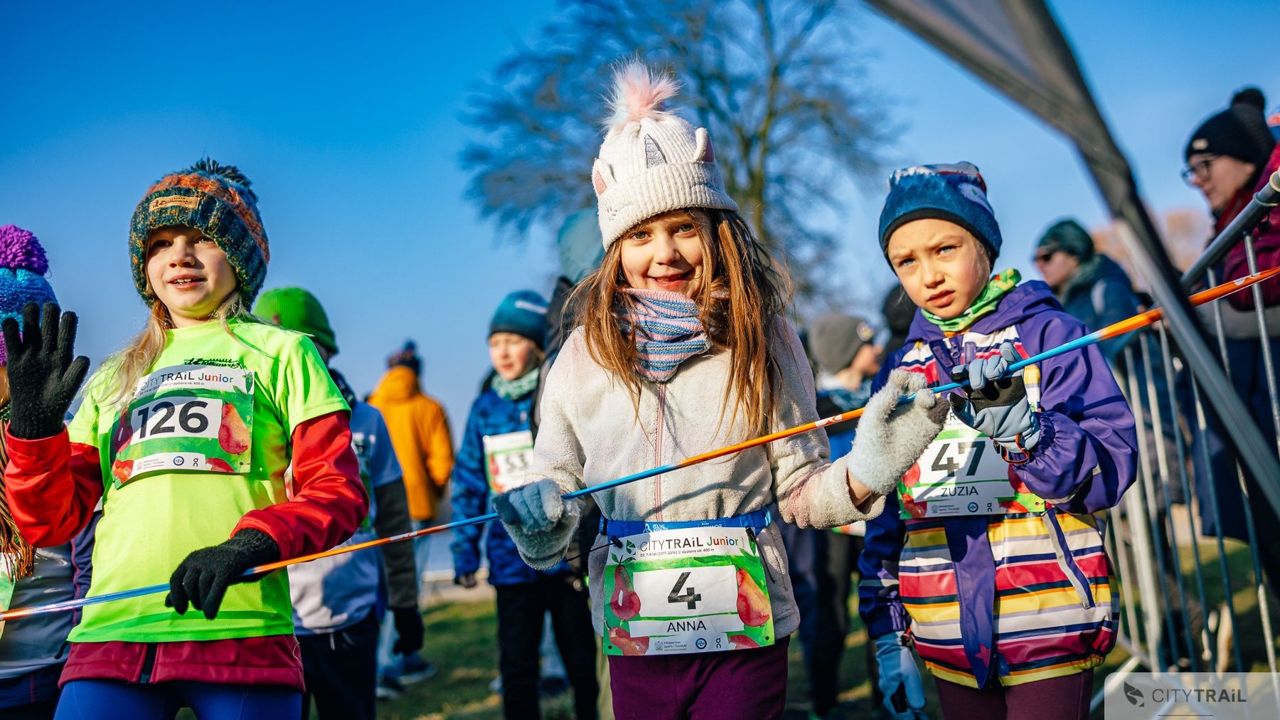 Bieg City Trail w Poznaniu, fot. Andrzej Olszanowski - dzieci z numerami startowymi stojące przy trasie biegu. Na pierwszym planie dziewczynka z beżową czapką i bluzą, zaś po bokach dziewczynki w zielonej i fiolettowej odzieży