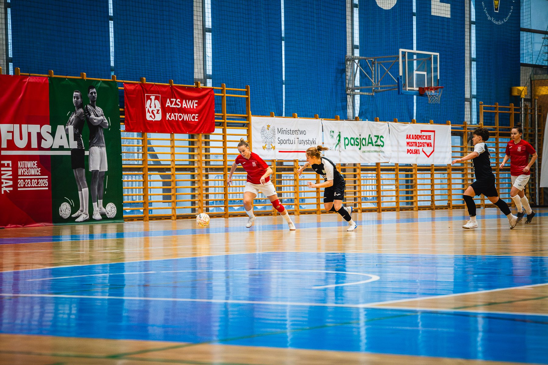 Finał Akademickich Mistrzostw Polski kobiet w futsalu z udziałem UAM Poznań, fot. Kamil Rojek - na sali grają po 2 zawodniczki w czerwono-białych i czarno-białych strojach. Przy piłce zawodniczka w czerwono-białym stroju. W tle drabinki i czerwony baner AZS AWF Katowice, a także biały Ministerstwa Sportu i Turystyki, Akademickich Mistrzostw Polski, oraz AZS. - grafika artykułu