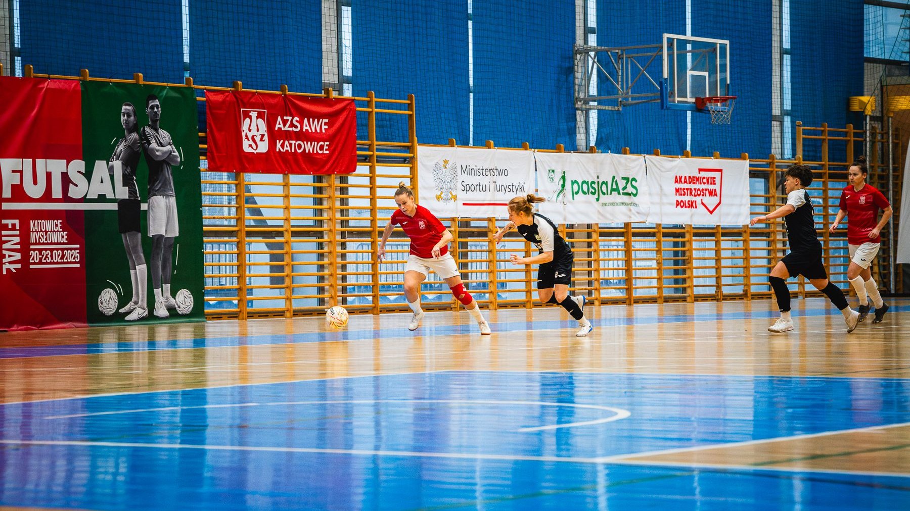 Finał Akademickich Mistrzostw Polski kobiet w futsalu z udziałem UAM Poznań, fot. Kamil Rojek - na sali grają po 2 zawodniczki w czerwono-białych i czarno-białych strojach. Przy piłce zawodniczka w czerwono-białym stroju. W tle drabinki i czerwony baner AZS AWF Katowice, a także biały Ministerstwa Sportu i Turystyki, Akademickich Mistrzostw Polski, oraz AZS.