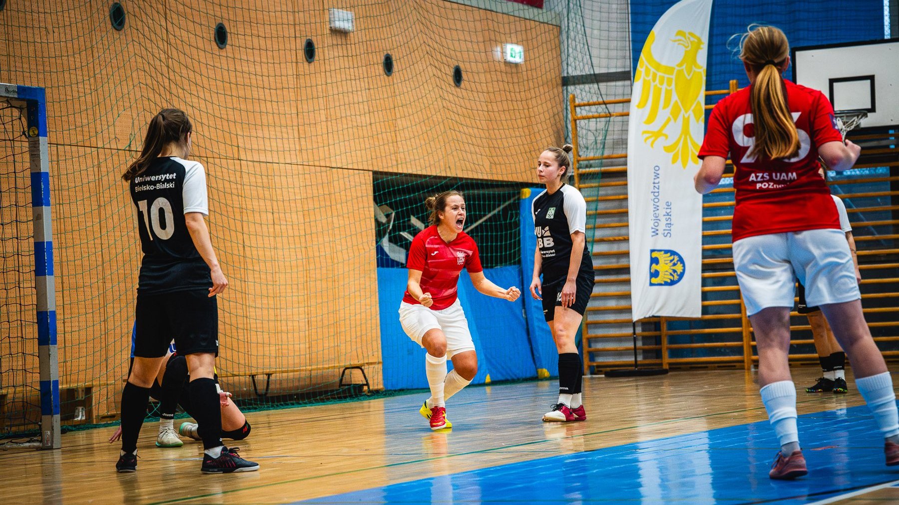 Finał Akademickich Mistrzostw Polski kobiet w futsalu z udziałem UAM Poznań, fot. Kamil Rojek - w sali 4 zawodniczki - po 2 w czerwono-białych i czarno-białych strojach. Cieszy się zawodniczka w czerwono-białym stroju. W tle baner Województwa Śląskiego