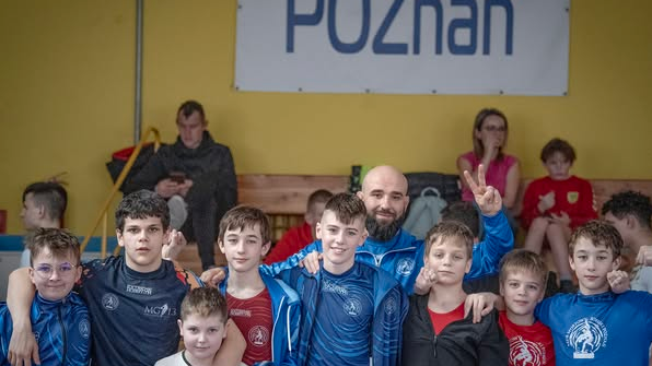 Otwarte Mistrzostwa Poznania w zapasach/Memoriał Bogdana Brody, fot. Maciej Kula - wspólne zdjęcie uczestników zawodów z trenerem. W tle widać kibiców na trybunach oraz baner Poznania na żółtej ścianie