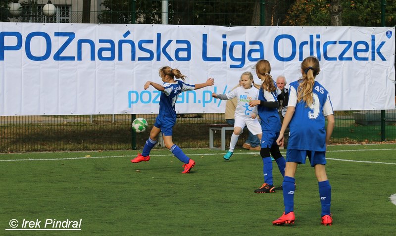 Poznańska Liga Orliczek, fot. Irek Pindral - na boisku 4 zawodniczki grają w piłkę nożna, jedna w białym stroju oraz 3 w niebieskich. W tle widać siedzących na ławce 2 opiekunów, a także baner z nazwą imprezy na białym tle - grafika artykułu