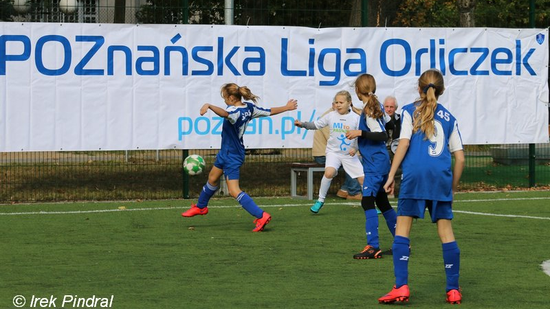 Poznańska Liga Orliczek, fot. Irek Pindral - na boisku 4 zawodniczki grają w piłkę nożna, jedna w białym stroju oraz 3 w niebieskich. W tle widać siedzących na ławce 2 opiekunów, a także baner z nazwą imprezy na białym tle