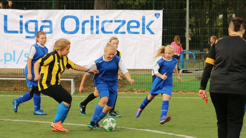 Poznańska Liga Orliczek, fot. Irek Pindral - na boisku widać 5 dziewczynek grających w piłkę nożna - 3 w niebieskich strojach oraz 2 w żółto-czarnych. Obok widać sędziującą kobietę, a w tle bawiące się dzieci, pustą ławkę oraz baner z nazwą imprezy na białym tle