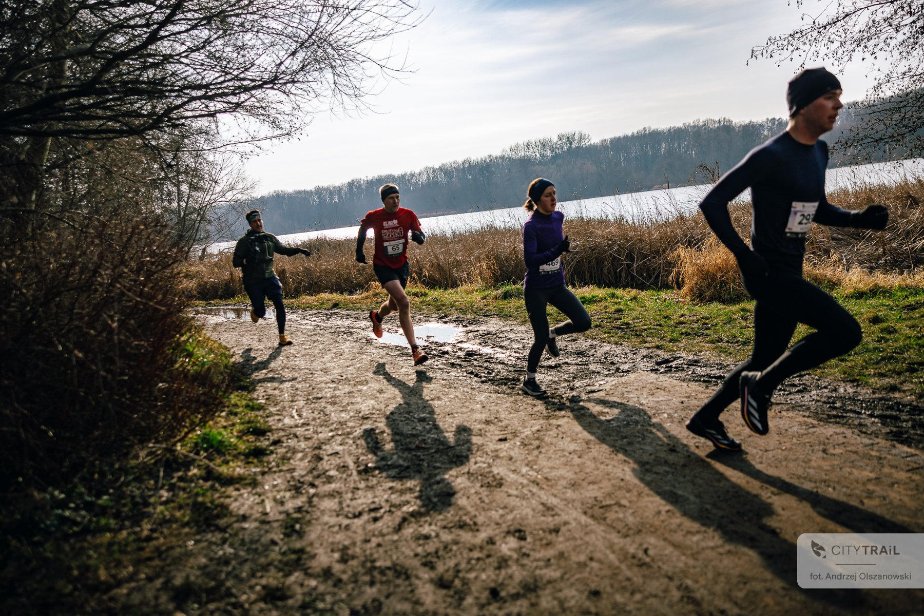 Bieg City Trail, fot. Andrzej Olszanowski - 3 zawodników i zawodniczka biegną w słonecznej aurze po grząskim terenie. W tle las oraz jezioro - grafika artykułu